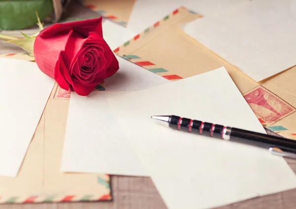 Vintage envelopes, red rose and sheets of paper scattered on the wooden table for writing romantic letters.
