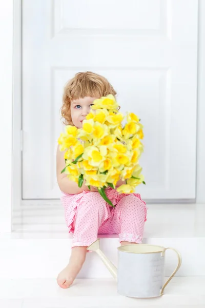 Little cute girl in a pink dress sitting on a white rustic porch and hides her face behind a bouquet of yellow garden flowers.