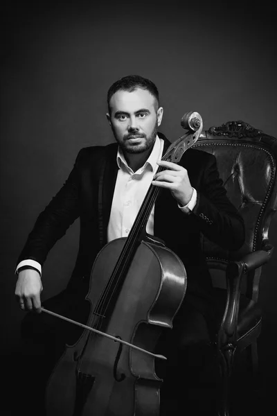 Portrait of young man in black suit sitting on luxury chair with cello