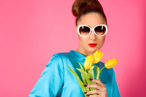 Portrait of young beautiful woman in blue silk dress and white sunglasses with yellow tulips ot the pink background