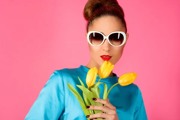 Portrait of young beautiful woman in blue silk dress and white sunglasses with yellow tulips ot the pink background