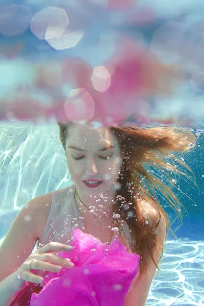 Young beautiful white woman in dress with pink flower underwater in the swimming pool