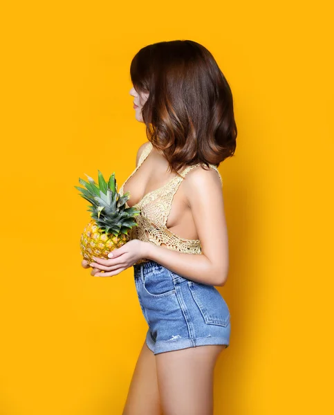 Beautiful sexy young woman in a short denim chert and  transparent top is on a yellow background sideways and  holding a pineapple in her hands round ass perfect body,  body oil with bronze skin bikini
