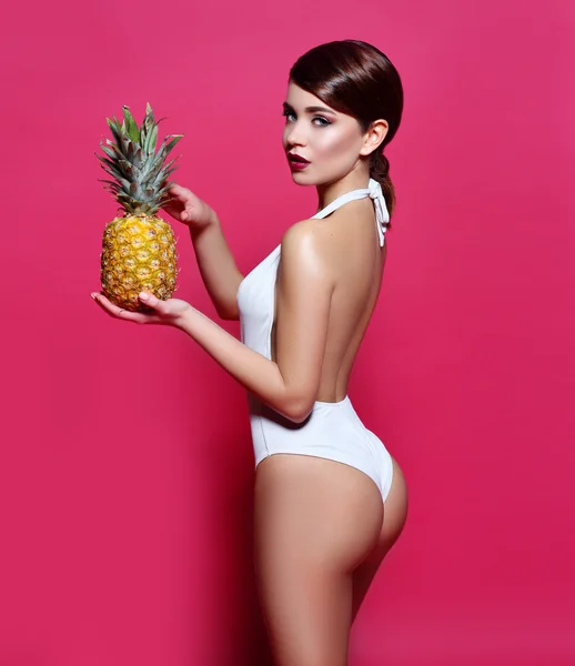 Beautiful sexy young girl standing on a pink background with  magazine  cover with perfect body with bronze tanned skin holding a pineapple  cocktail smiles a lovely smile perfect photos for advertising
