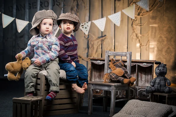 Two little boys in caps sitting on boxes in the studio around soft toys bears