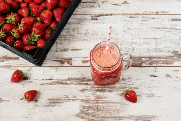 Fresh juice, shake, milkshake of strawberries in a mason jar with a straw. Pile of juicy ripe organic fresh strawberries in a black crate. Strawberry fresh drink. Light rustic wooden background