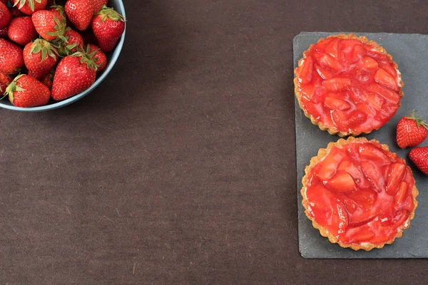 Chocolate tart, tartalette with white chocolate and mascarpone cream, fresh strawberries on top. Large blue bowl full of strawberries. Black background. Empty space