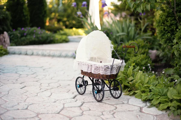Doll\'s pram. Vintage doll stroller placed on the stone walkway, alley in a beautiful garden with flowers and trees around. Retro cart dolls made of rattan and white lace