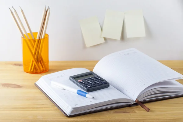 Notepad with pen container with pencils, calculator are on a wooden table. On the wall near the table glued paper for notes.