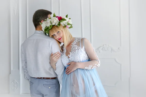 Beautiful pregnant woman in light blue dress with flower diadem and ger husband in white room