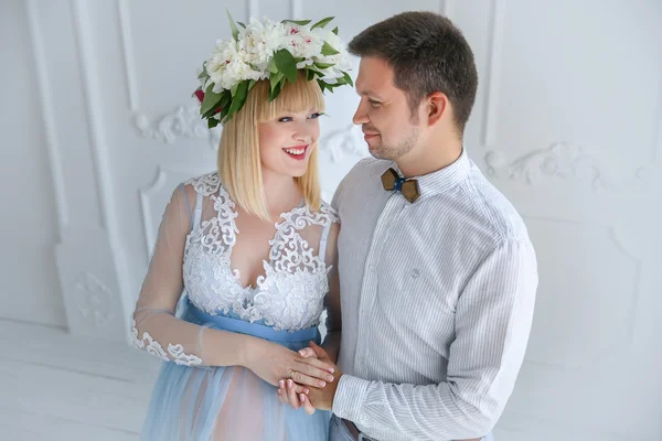 Beautiful pregnant woman in light blue dress with flower diadem and ger husband in white room