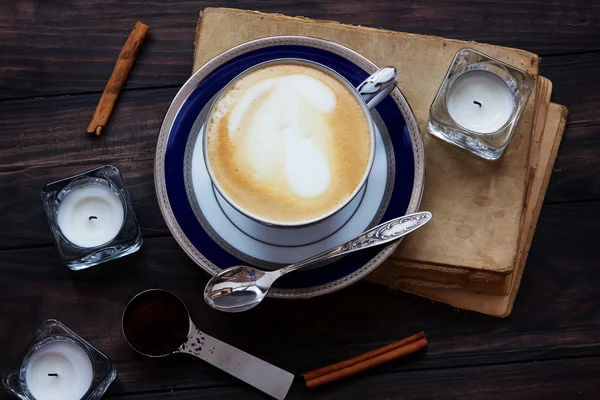 Coffee composition with old book, white tea candles, silver spoon, ground coffee and cinnamon on dark wooden background.