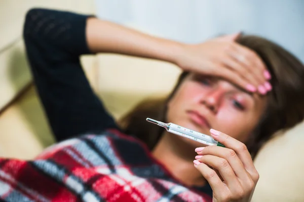 Sick woman lying on sofa under wool blanket and looking at thermometer