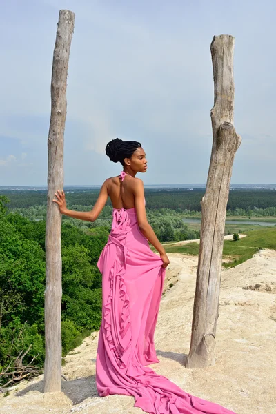 African American girl with dreadlocks, in a pink dress, standing with her back on a high hill
