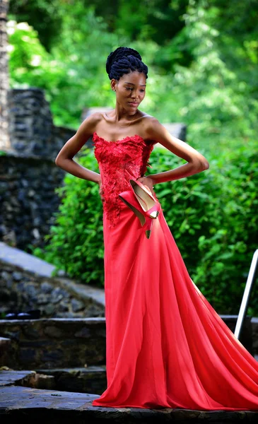 African American girl in a red dress, with dreadlocks, with red shoes in hand, posing in the summer in the Park on a background of green plants
