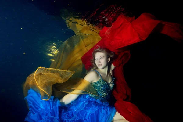 Beautiful girl dancing underwater with red and orange cloth in blue dress on a black background and looks away. Portrait. Horizontal orientation.