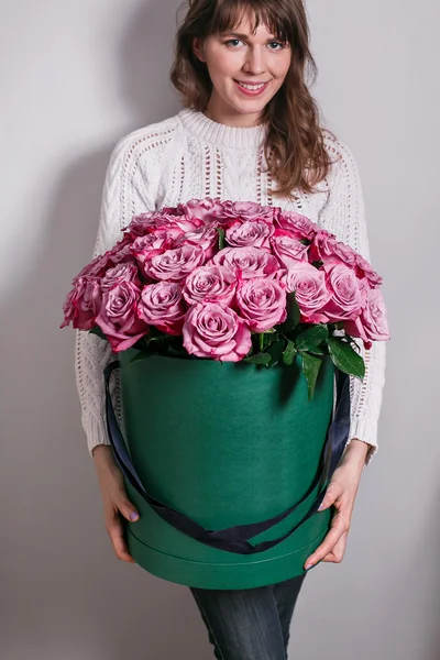 Nice young blonde girl. smelling flowers holding purple roses bouquet in hat box against the plastered wall, wearing jeans knit sweater.