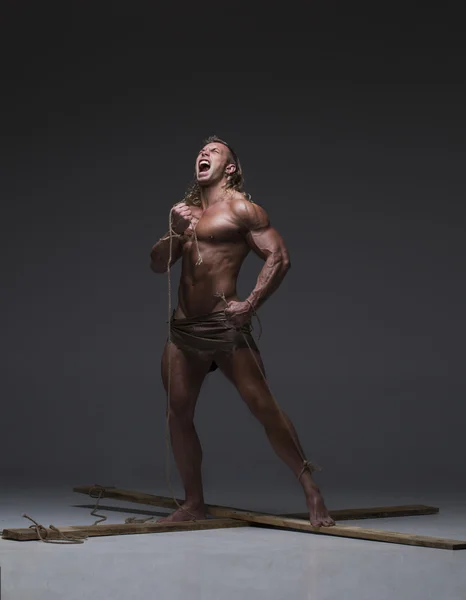 Screaming naked muscular man, hands tied rope to wooden beams