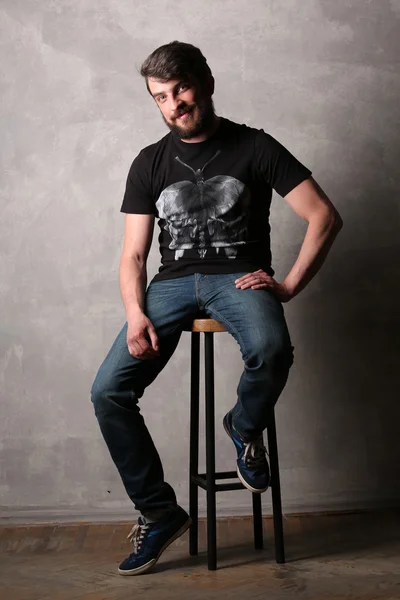 Bearded man in black T-shirt with sitting on a bar stool. Gray