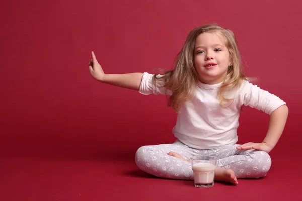 Girl in pajamas with glass of milk showing finger. Red background