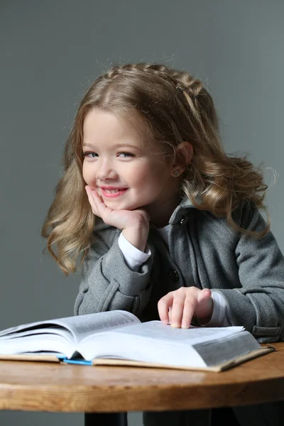 Little blonde sitting at the table with a book. Close up. Gray background