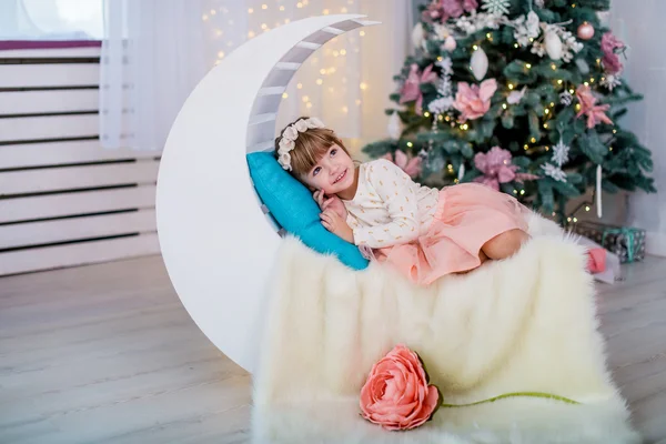 Happy little girl lying under the tree on decorative months against the background of Christmas lights