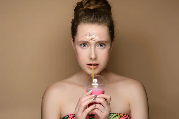 Portrait of young beautiful girl with milk shake coctail in hand on the gold background. Nude makeup with tinsel. Concept of the beauty.