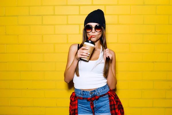 Beautiful young sexy girl drink coffee, smiling and posing near yellow wall background in sunglasses, red plaid shirt.