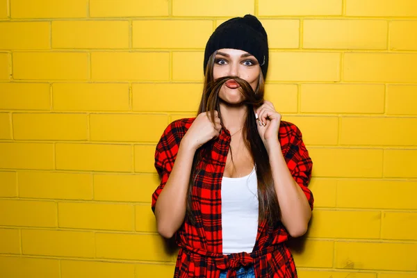 Beautiful young sexy hipster girl posing, smiling, do fake mustache near urban yellow wall background in red plaid shirt, shorts, hat.