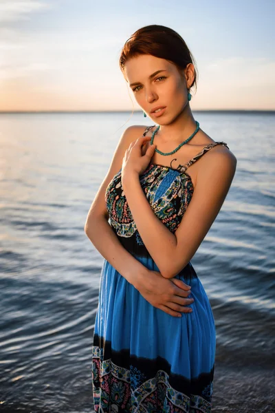Young beautiful girl in blue dress posing at sea coast against blue sky at sunset