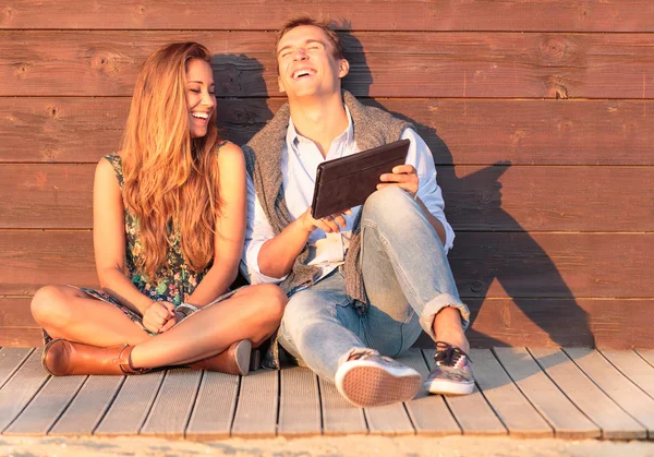 Cheerful guy with girl laugh about video on the tablet. Best friends having fun at the beach with social media and funny contents. Friendship of young people during holiday life moment at sunset.