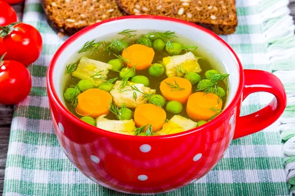 Chicken soup in a red bowl with carrots, peas and greens on a na