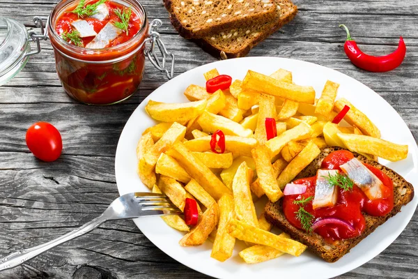 Potato Fries with pieces of chili pepper on the white dish and Slices of marinated Norwegian herring in tomato sauce on the pieces of rye bread, the glass jar with  delicious herring with dill, top view