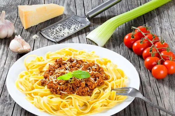 Bolognese ragout with italian pasta on a white plate, decorated with basil leaves, authentic recipe, wooden background with celery, garlic, cherry tomatoes and parmesan cheese, full focus, close-up