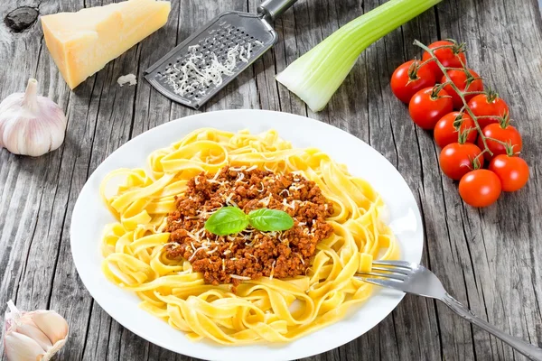 Bolognese ragout with italian pasta on a white plate, decorated with basil leaves, authentic recipe, wooden background with celery, garlic, cherry tomatoes and parmesan cheese, full focus, close-up