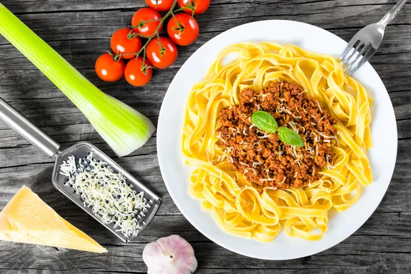 Bolognese ragout with italian pasta on a white plate, decorated with basil leaves, authentic recipe, wooden background with celery, garlic, cherry tomatoes and parmesan cheese, full focus, close-up