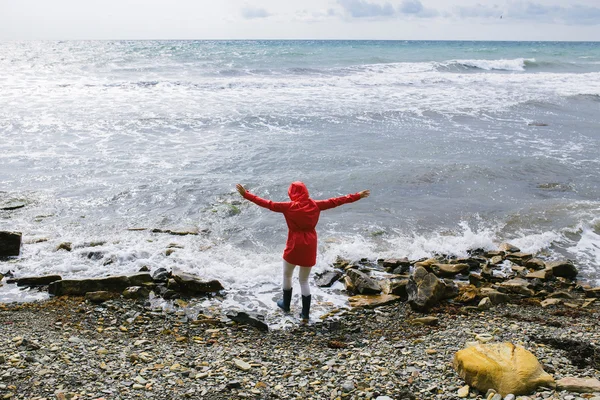 Young woman in a red raincoat and blue rubber boots comes into sea water with raised arms
