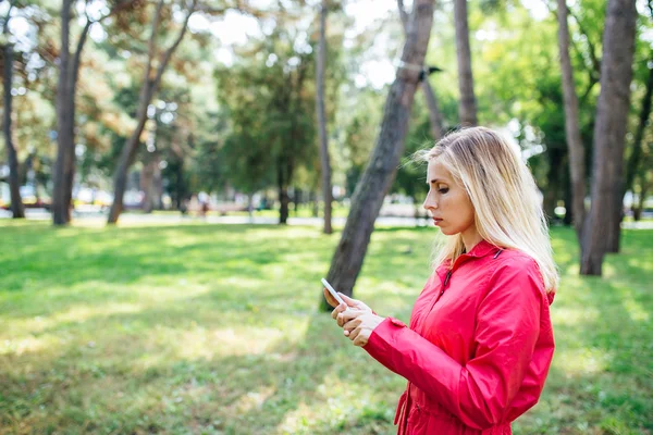 Young blonde woman in a pink jacket sending a text message from her cell phone in park. Woman using smart phone outdoor