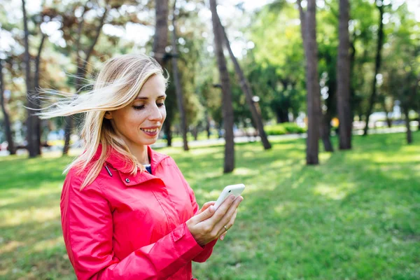 Young blonde woman in a pink using smart phone in park. Woman reading text message on the phone outdoor.