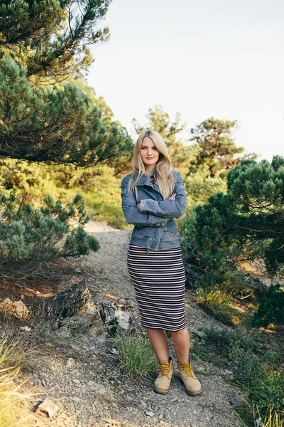 Portrait of a beautiful young blonde woman in denim jacket, striped dress and yellow boots standing with folded hands outdoor