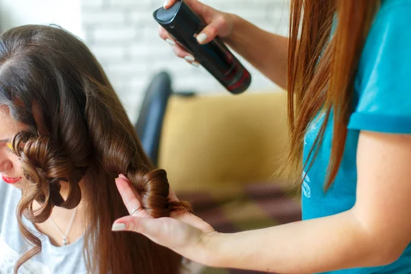 Closeup of hairdresser doing the styling for a festive evening of wedding hairstyles woman with long black hair. Hairdresser makes curles using hairspray. Fashion concept.