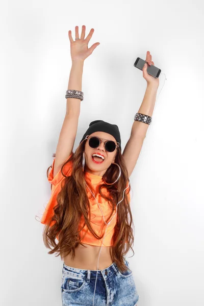 Beautiful woman in sunglasses wearing in black hat and orange T-shirt listening music and dancing near white wall, holding a cell phone in hand, hands raised up