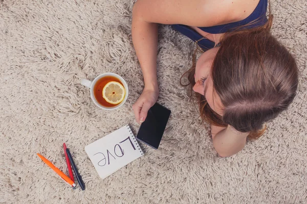 Beautiful girl lying on the carpet at home and reading sms, next to her cup of tea with lemon stand, top view
