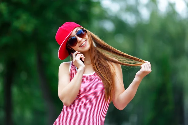 Fashion city portrait of stylish hipster woman talking mobile phone, red striped dress, red cap and sneakers, makeup, sunglasses, long brunette hairs, walking alone at weekend, enjoy vacation in