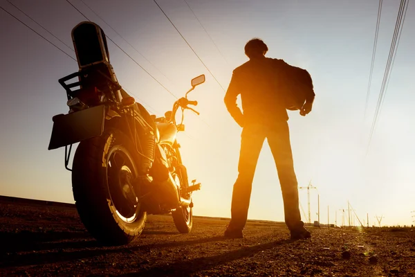 Biker with motorbike stands on sunset backdrop