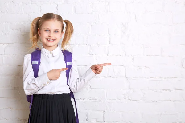 Cute smiling schoolgirl in uniform standing on light background and showing thumbs to the side.
