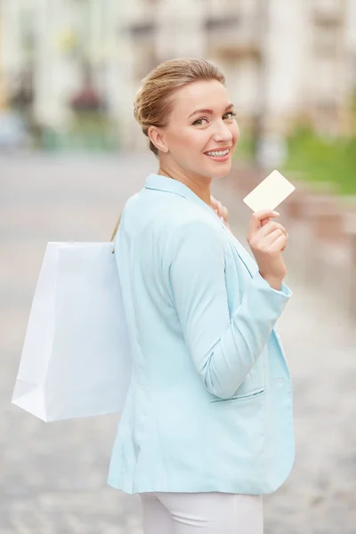 Beautiful woman standing half-turned holding bag for shopping and showing credit card on the street city.