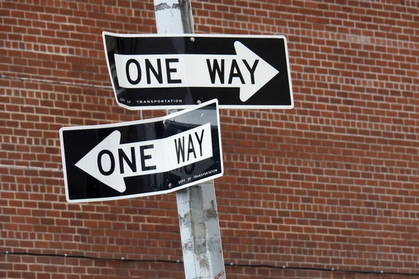 Two One Way Signs pointing in two different directions in front of a red brick wall