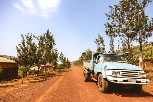 The light blue old truck carrying the workers on the road surrounded by trees.