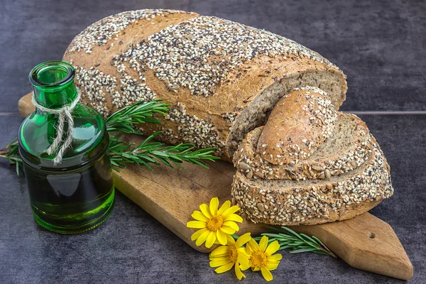 Whole wheat bread and rye, sprinkled with sunflower seeds, poppy , sesame seeds, sliced on the board, next to a jar of olive oil, rosemary and yellow daisies on a dark background